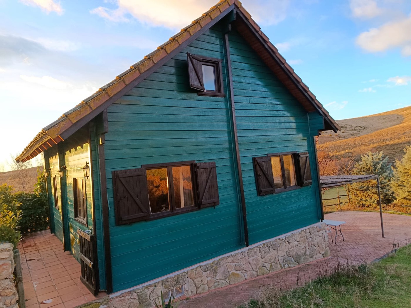 Casa de madera verde de The Forge al atardecer, con tejado de teja y base de piedra, en la dehesa de El Molar, Madrid